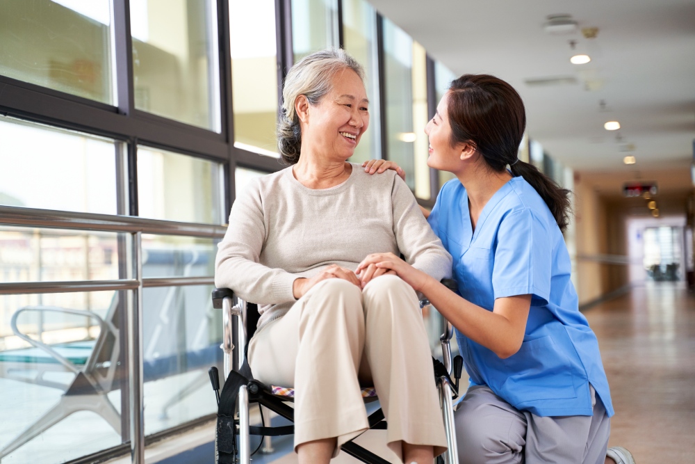 Smiling elderly woman in wheelchair interacting with compassionate healthcare professional in a clinical setting, illustrating support and independence in daily living activities.